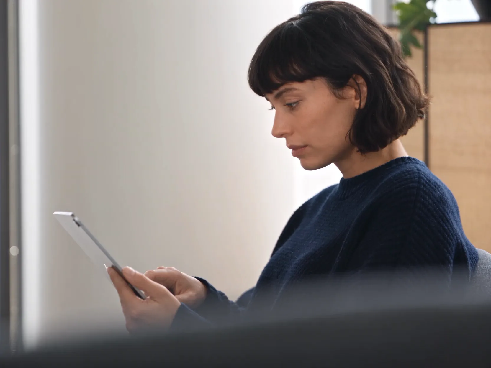A data analyst with a dark bob in a navy knit, seated in a bright lounge, reading her iPad and smiling at what she finds.