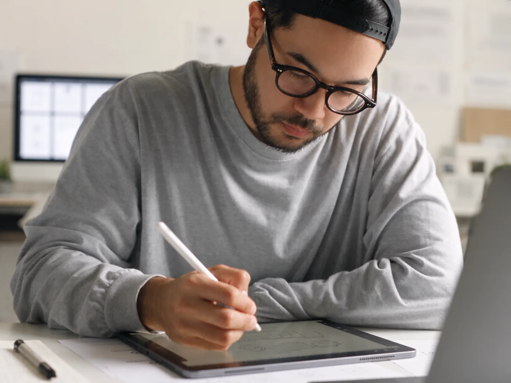 A designer in glasses and a cap drawing on an iPad with a stylus at a bright studio desk.